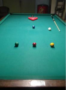 A snooker table has 14 red balls and numbered balls 2-7, each ball having a specific place on the table. Although it isn’t shown, the yellow 2 ball, red 4 ball and green 3 ball all sit on a line that is part of a half circle. This is the break circle. The cue ball is placed here when the shooter breaks the rack and also after a scratch. Further down, you see the blue 5 ball, and in the middle of the rack is a pink 6 ball. In the very back is the black 7 ball. As you can see, the pockets are much smaller, and the table longer and wider, than a standard billiards table. 